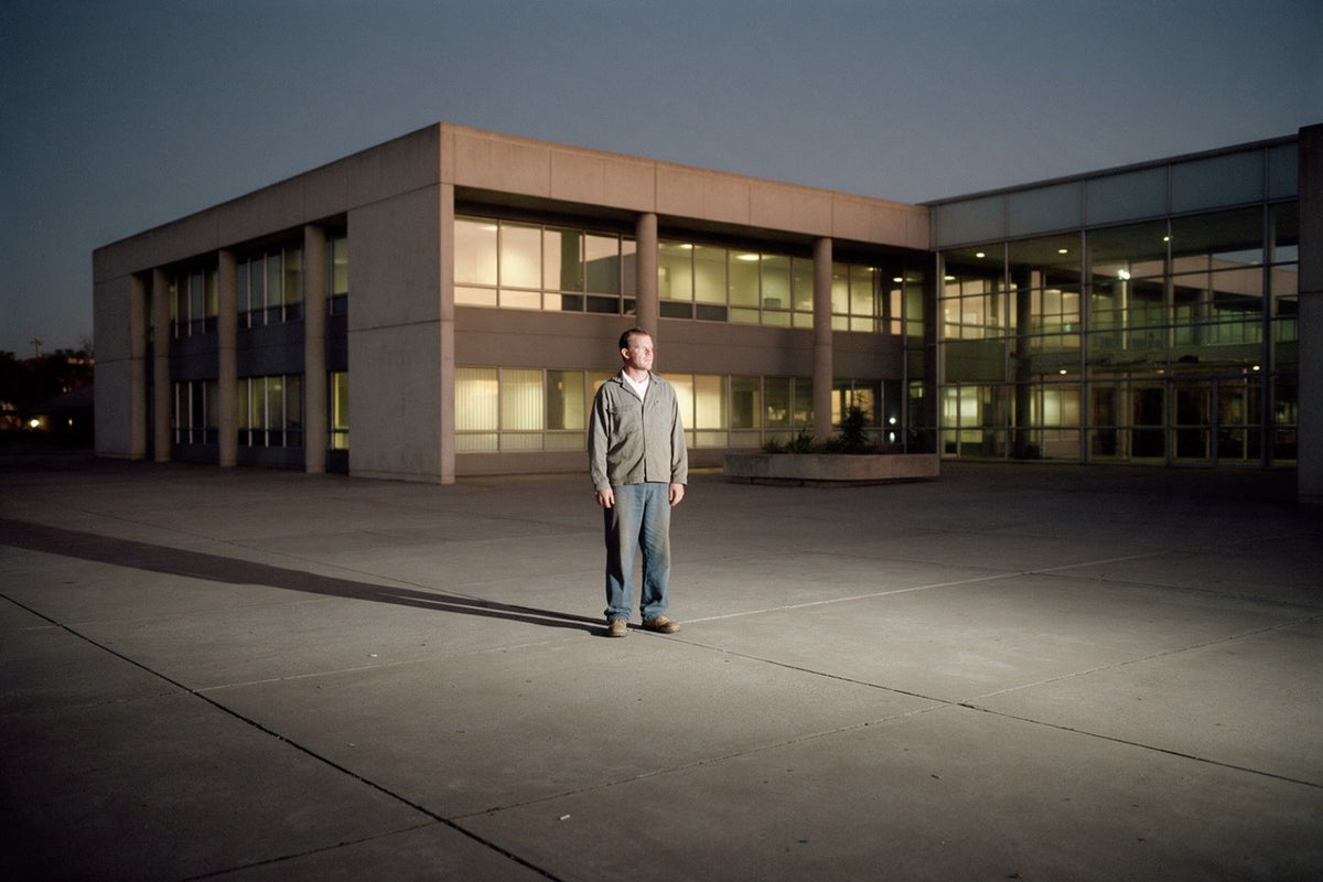 A man standing alone in an empty office forecourt at dusk